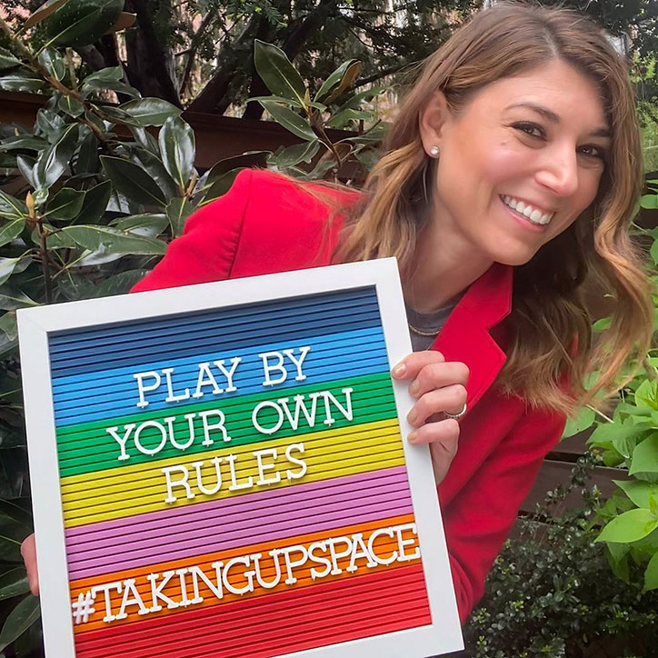 Author Alyson Gerber wears a red pantsuit and holds up a rainbow sign stating "Play By Your Own Rules #TakingUpSpace"
