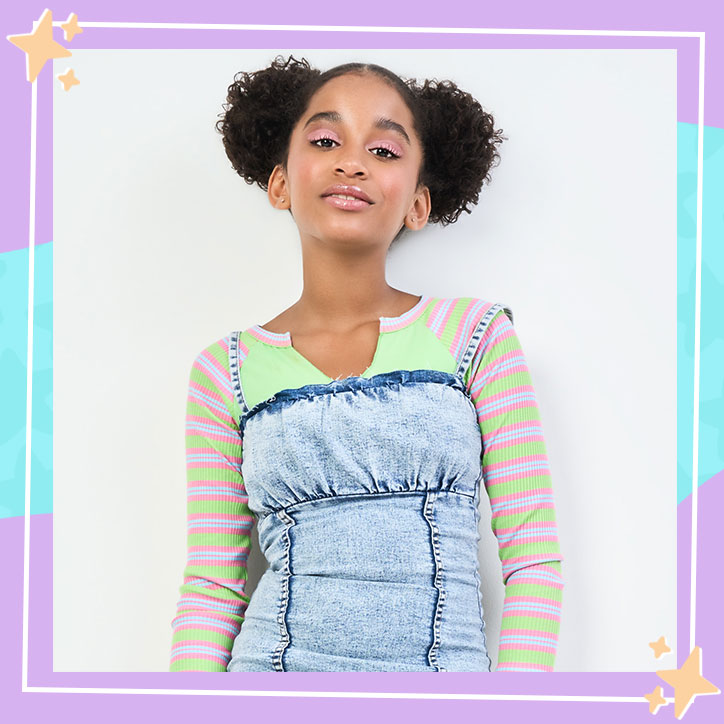 Actress Jordyn Curet poses against a blank wall, wearing a colorful striped shirt, denim dress, and her hair up in space buns