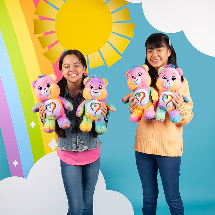 Two tween girls standing in front of a rainbow backdrop holding up Togetherness Bear Care Bears plush toys