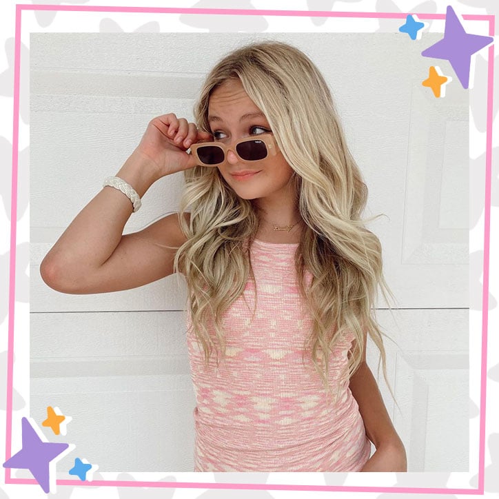 Pressley Hosbach poses in front of a wall in a pink dress, tipping her sunglasses