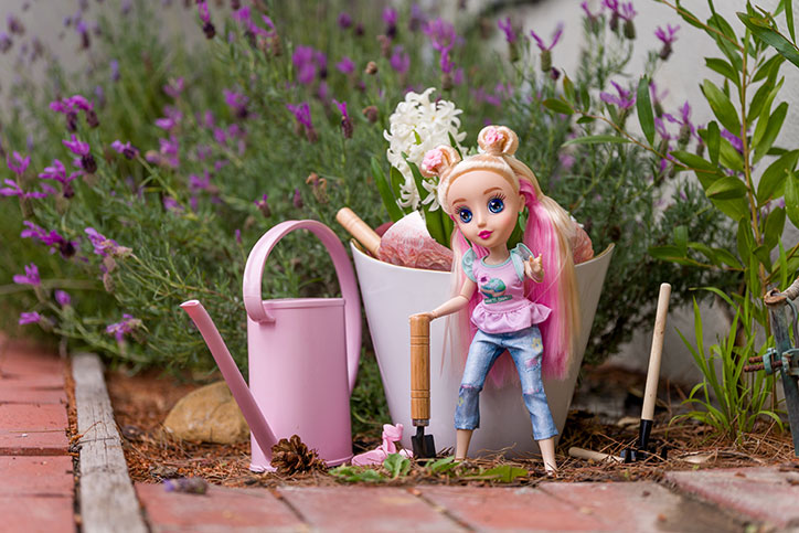 Brianna of the B-Kind Dolls standing in a garden next to a pink watering can and bucket of gardening tools.