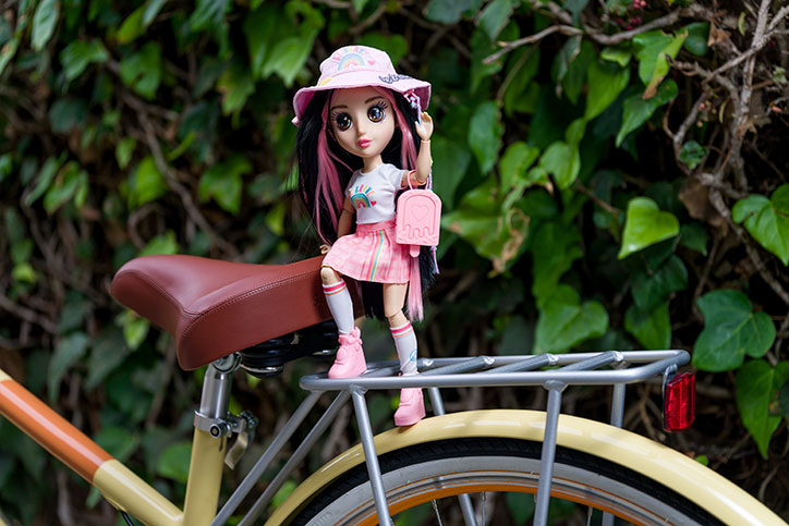 Nora from the B-Kind Dolls sits on the rack of a bicycle