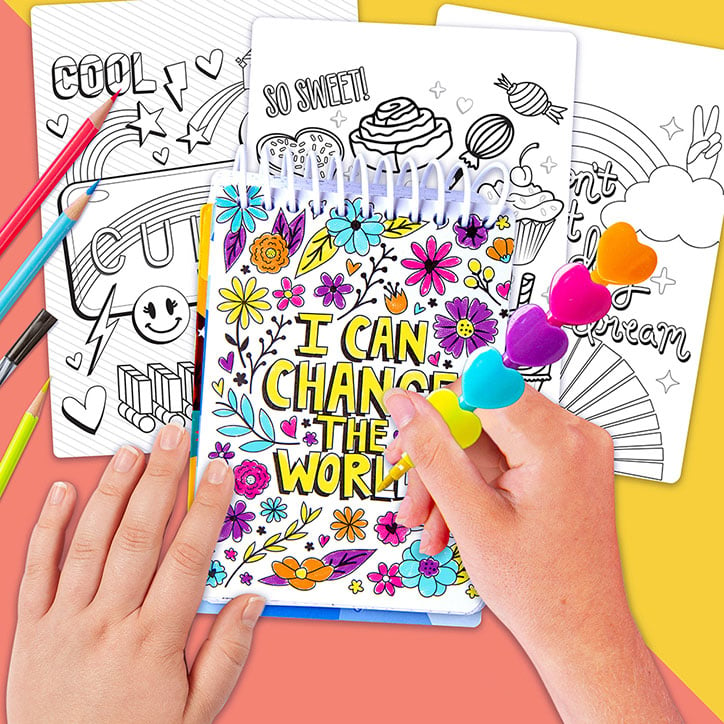 Flat lay of a girl using the included heart shaped crayon, coloring a quote graphic that says "I Can Change the World" from the Fashion Angels Positivity Doodles Activity Set