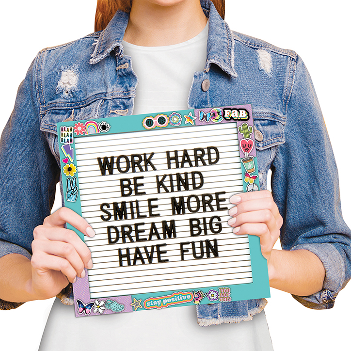 Tween girl in a denim jacket holding up the letter board found in the Fashion Angels Letter Board Design Kit. Letter board is covered in stickers around the frame and the message says "Work Hard, Be Kind, Smile More, Dream Big, Have Fun"