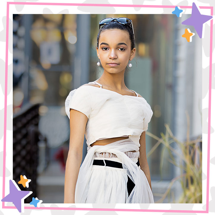 Kherrington Briggs poses outdoors in an elegant white crop that wraps and ties around her waist and a belted white skirt and pearl earrings