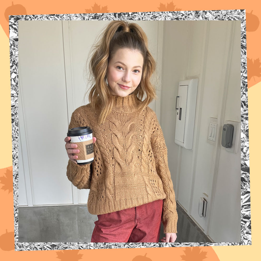 Lilo Baier poses in a tan cable knit sweater and salmon pants while holding a pumpkin spice latte