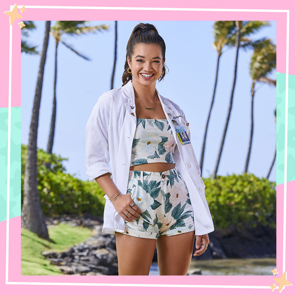 Peyton Elizabeth Lee poses in character as Lahela Kamealoha in front of palm trees in her doctor coat and floral outfit