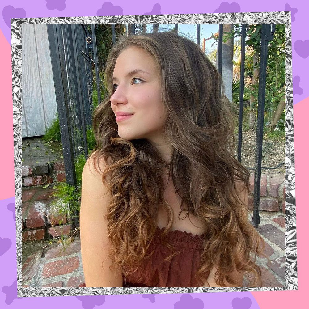 Chiara D'Ambrosio poses outside in front of an iron fence with her head turned to the left. She is wearing a brown ruffly tanktop and her long wavy hair covers her shoulders.