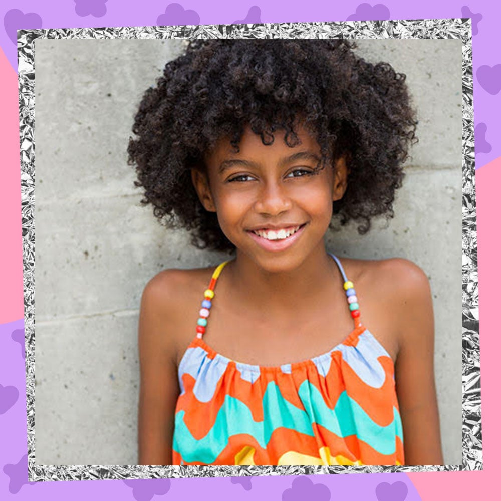 Hannah Love Jones poses in front of a cement wall, smiling, in a wavy rainbow tanktop with her natural afro-style hair
