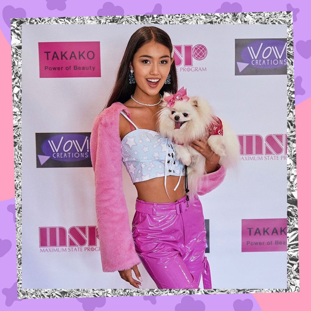 Kylin Kalani poses in front of a step and repeat while holding a fluffy white pomeranian. She is wearing a blue cropped tank, pink pleather pants, and a fluffy pink sweater