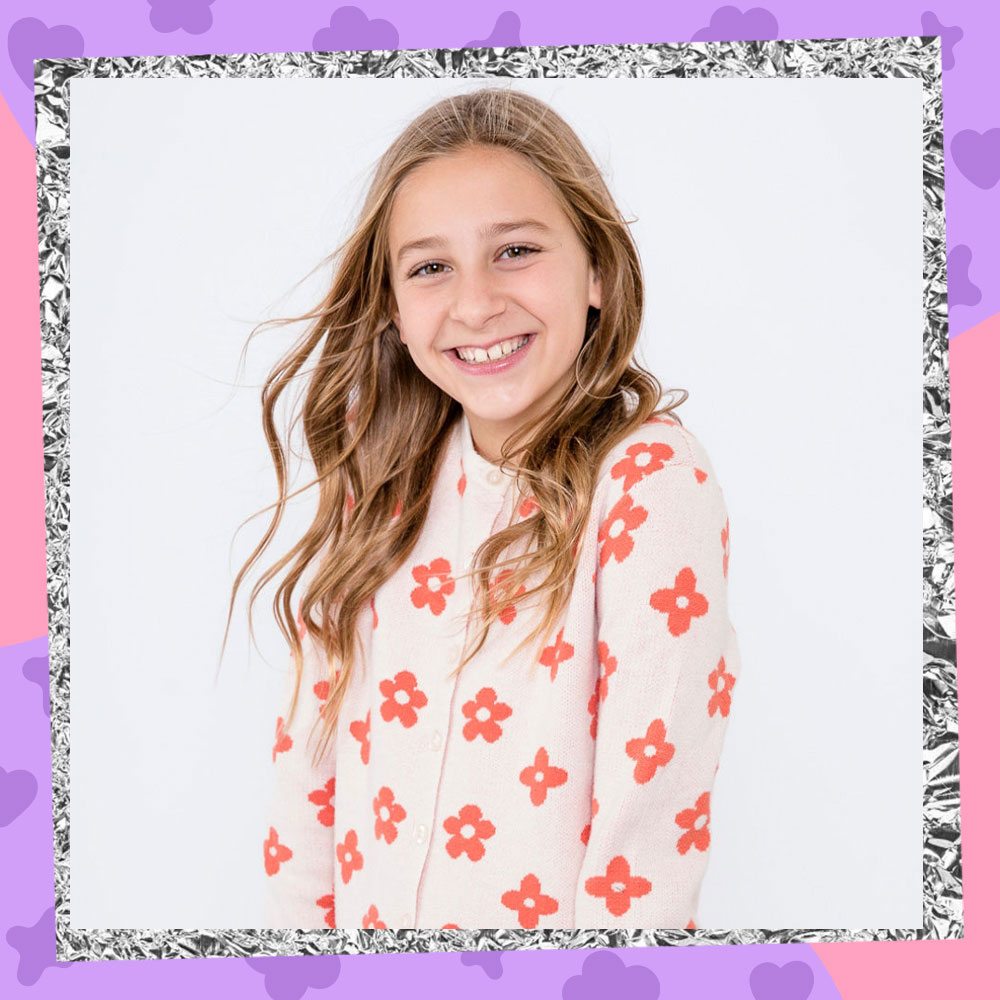 Photoshoot image of Sky Alexis with her hands in her pockets, smiling, while wearing a white cardigan with retro orange flowers