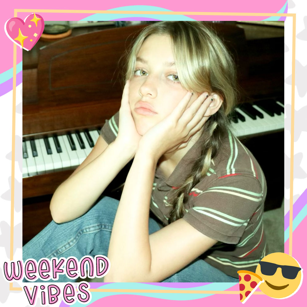 Ava Grace poses in front of a piano with her hands on her cheeks while wearing jeans, a brown striped shirt, and pigtail braids