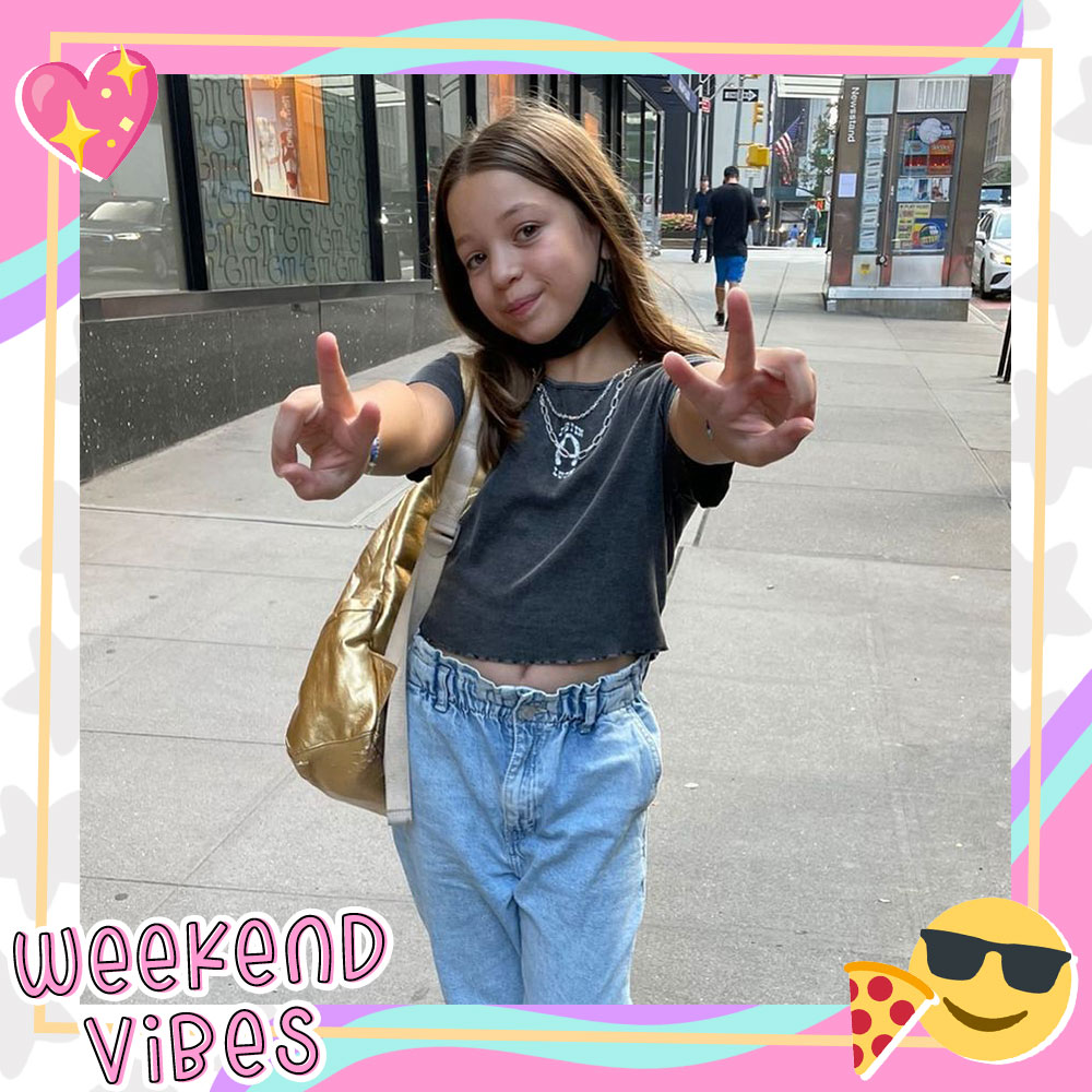 Dalya Knapp poses on the street holding up two peace signs. She's wearing a grey tshirt, light blue baggy jeans, a metallic gold purse, and lots of necklaces.