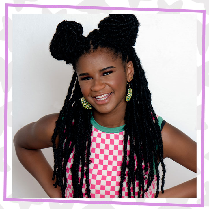 Picture of Marley Dias posed with her hands on her hips in front of a blank wall. Her braided hair is half up in buns, half hanging down and she is smiling and wearing a pink and white checkered tank with green trim.