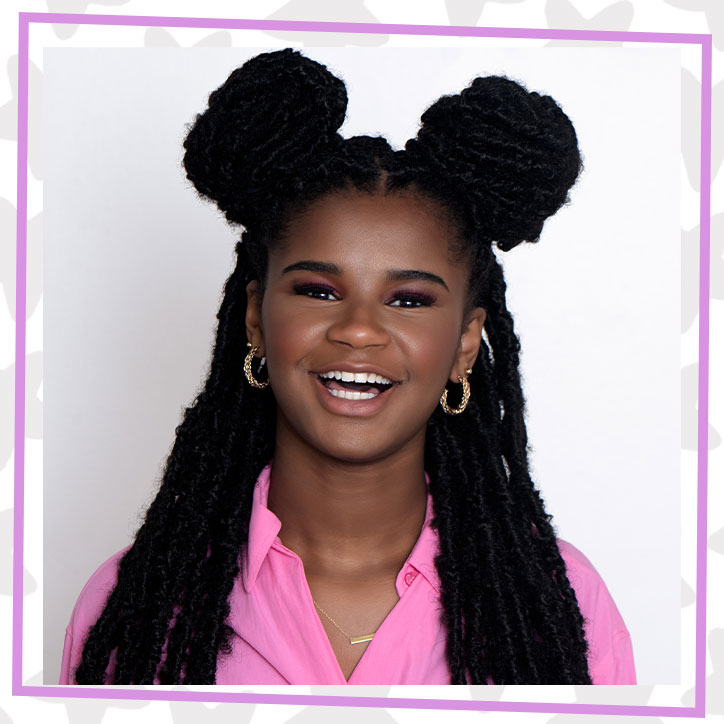 Close up of Marley Dias smiling in a pink button down shirt. She has her braided hair half up in buns, half hanging down and is wearing gold hoop earings.