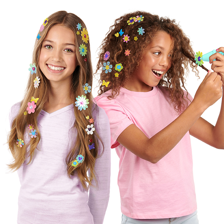 Two girls standing together using the Style Squad Hype Hair Floral Frenzy hair stamper. Each girl has dozens of colorful paper flowers in their hair.