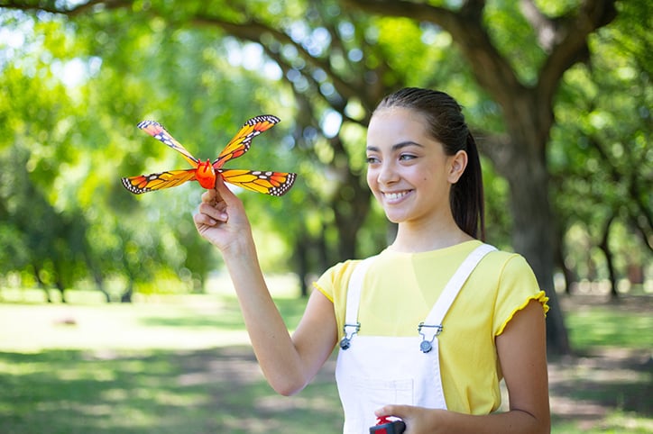 A teen girl stands in a park holding the ZING Go Go Bird Butterfly in one hand and the remote control in the other