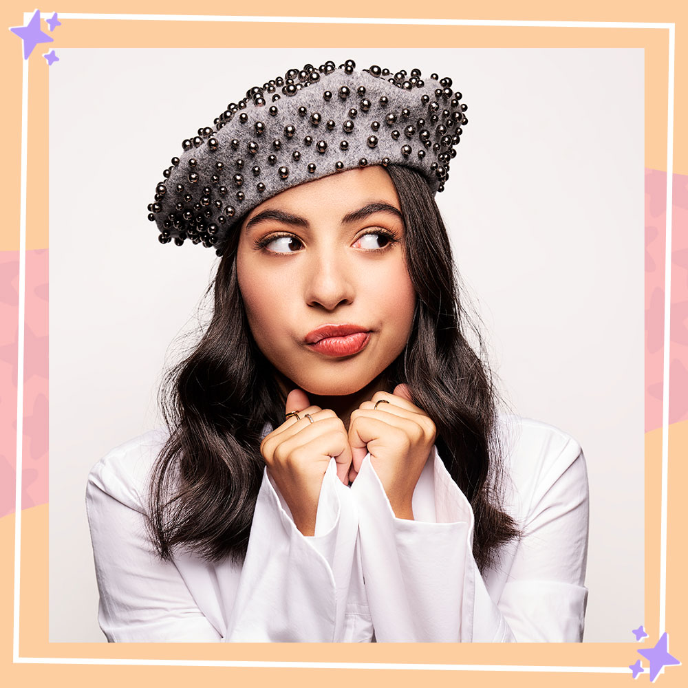 Kyndra Sanchez poses in front of a white backdrop while wearing a white button down shirt and a gray beret studded with pearls