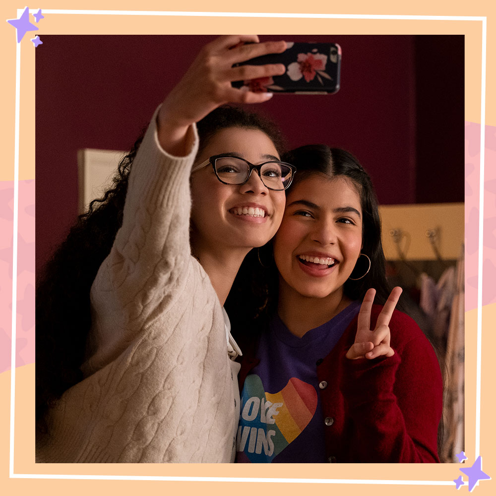 Photo from the set of The Baby-sitters Club featuring Malia Baker and Kyndra Sanchez in character as Mary-Anne and Dawn. They are taking a selfie, Malia is holding up a phone and Kyndra is posing with a peace sign