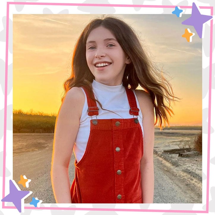 Kira McClure poses in front of a sunset on the beach while wearing burnt orange corduroy overalls and a white turtleneck tank