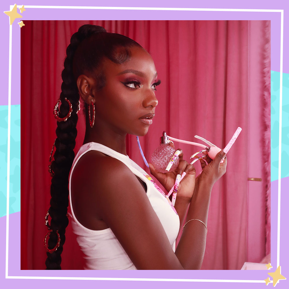 Kheris Rogers poses in front of a pink curtain and a vanity mirror. She is using an old fashioned spray perfume bottle.