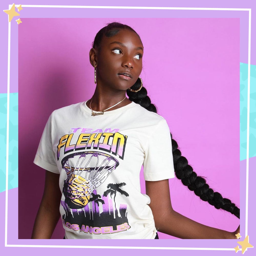 Kheris Rogers poses in front of a pink backdrop, holding her long braid to the side. She is wearing a shirt from her Flexin' in My Complexion clothing line.