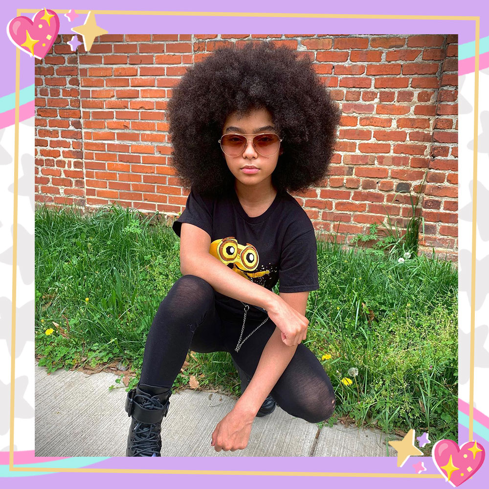Madelen Mills kneels in front of a grass patch and a brick wall. Her natural hair is styled in an afro and she is wearing aviator sunglasses, a black tee black leggings, and black combat boots
