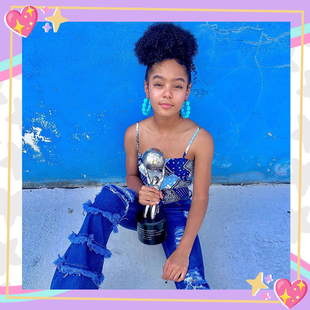 Madalen Mills sits in front of a blue concrete wall in ruffled denim jeans, a blue and white handkerchief style tank, and beaded blue hoop earrings. Her hair is up in a big bun and she is holding her NAACP Image Award for her role in Jingle Jangle