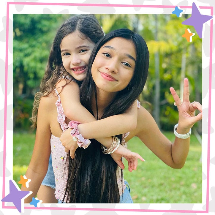 Jasmine and Bella Mir pose outdoors in a grassy park surrounded by trees. Jasmine has Bella on her back and is holding up a peace sign.