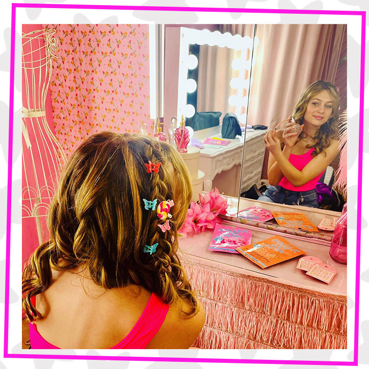 Lulu in the dressing room behind the scenes of the GLOTIVATION Rock Your GLO music video. She is looking into a mirror spraying on perfume and has colorful butterfly clips in her curled hair