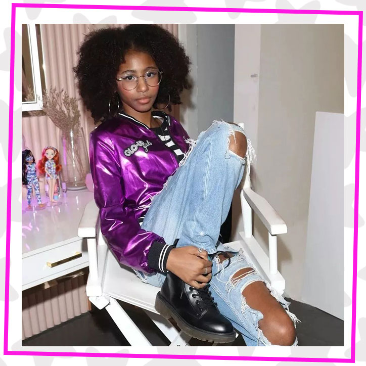 Saucy behind the scenes of the GLOTIVATION Rock Your GLO music video. She is sitting in a directors chair wearing a purple metallic GLOTIVATION bomber jacket, ripped jeans, and black combat boots.