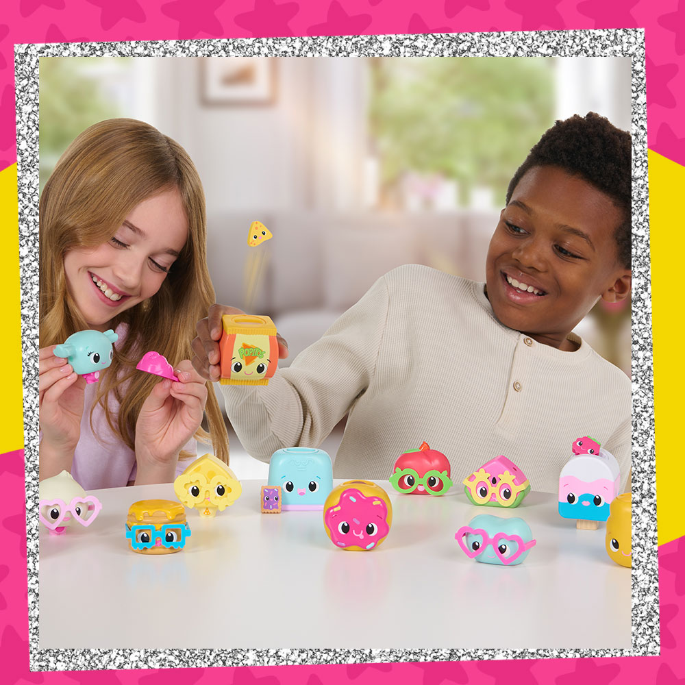 Lifestyle photo of two kids playing with an assortment of My Squishy Little Snack Pack figures including a bag of chips, a donut, a watermelon, and more.