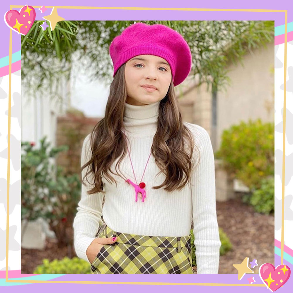 Madeleine McGraw poses in front of a house with greenery in the background. She is wearing a white turtleneck, a yellow plaid skirt, a pink baret, and a pink unicorn necklace.