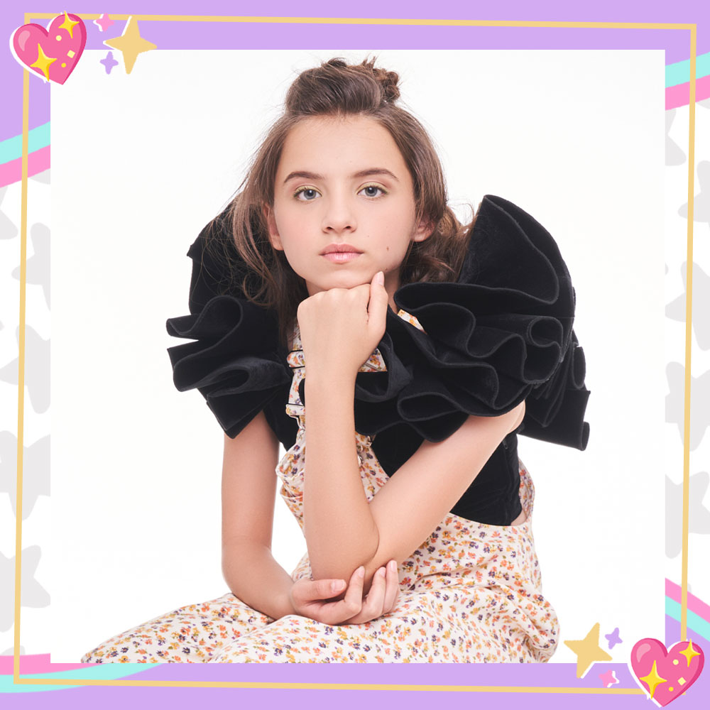 Madeleine McGraw is seated in front of a white backdrop, holding her hand to her chin. Her hair is in an updo and she is wearing a black dress top with oversized ruffled sleeves with floral overalls layered over it.