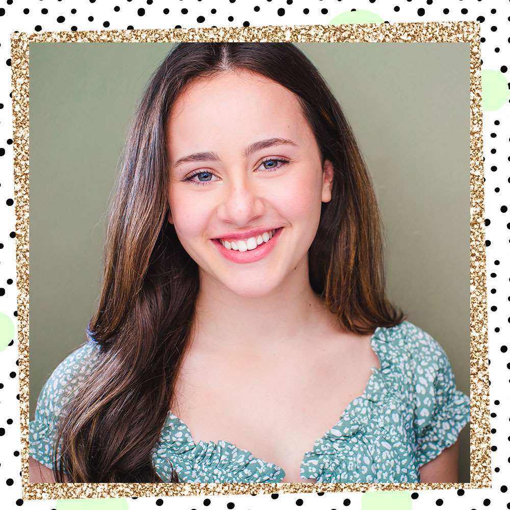 Abbie Grace Levi poses in front of a green backdrop. Her long hair is half behind her, half tossed over her right shoulder. She is wearing a floral blue top with ruffled puffy sleeves.