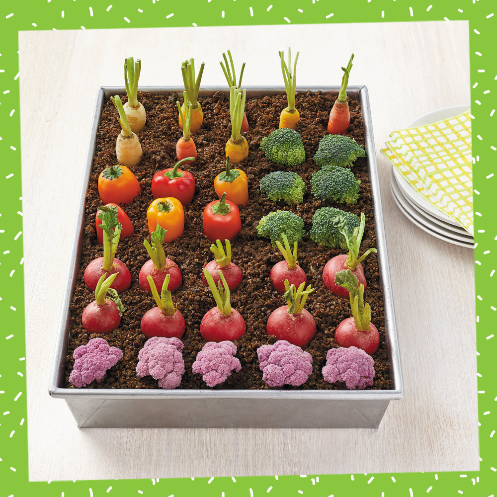 A rectangular baking dish sits on a kitchen table. It is filled with hummus and crumbled pumpernickel "dirt", and has vegetables laid out in rows to look like a garden plot. The recipe is the Garden Plot Veggie Dip from the Recipe-a-Day Kids Cookbook