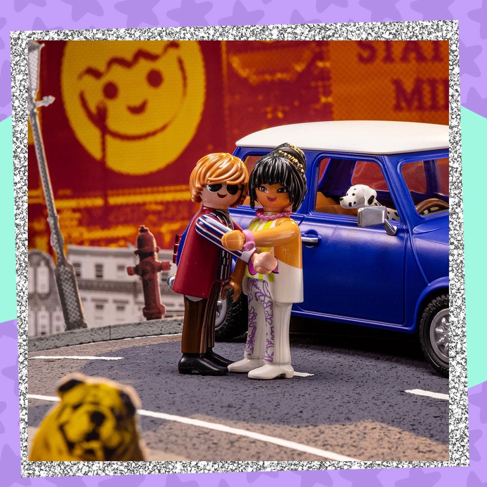 Lifestyle photo of the Playmobil Mini Cooper parked in a London-inspired alleyway. The man and woman figures are posed together in a hug while the Dalmatian peeks out the car window.