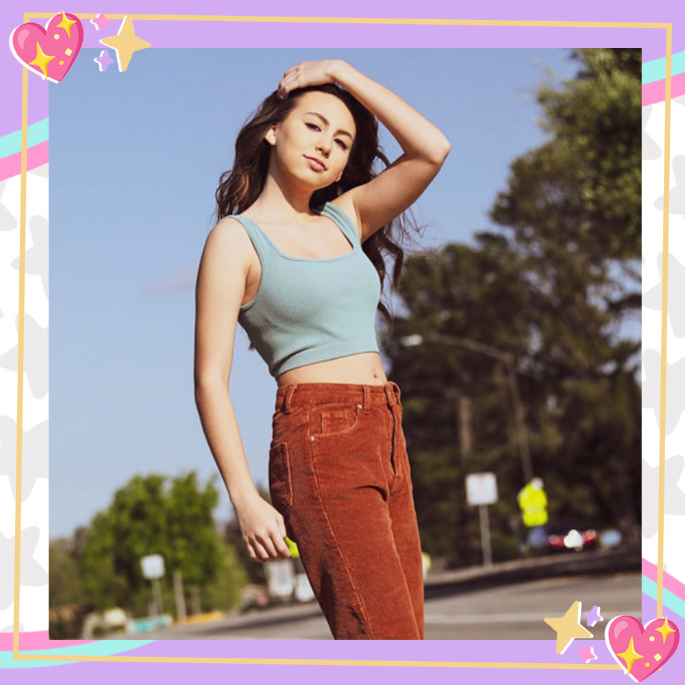 Image of Lola Raie skateboarding down the street. She is wearing a baby blue tanktop and burnt orange cordorouy jeans and casually pulling her hair back with her hand