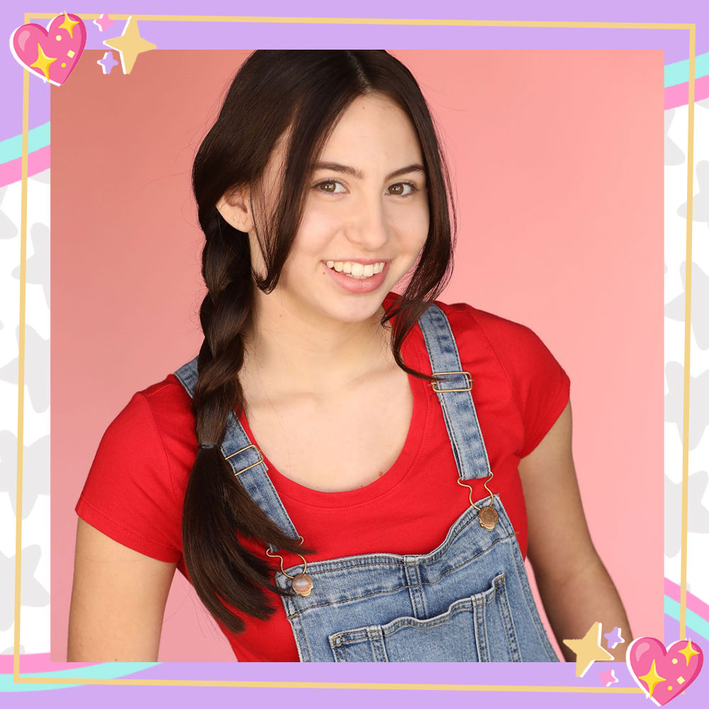 Lola Raie poses in front of a pink backdrop. She has her hair in braided pigtails and is wearing a red tshirt and denim overalls.