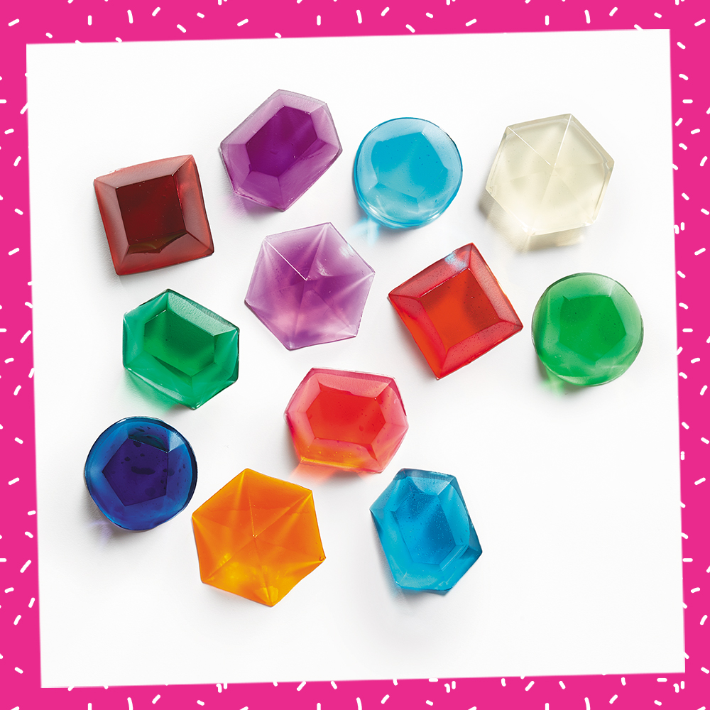 Lifestyle photo of the Gelatin Gems recipe from the Recipe-a-Day Kids Cookbook. A variety of colorful gemstones in different shapes sit spread out on a white backdrop.