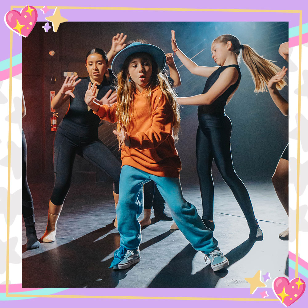 Mandy Corrente on the set of her "Super Duper" music video. She wears a baby blue hat, orange sweatshirt, baby blue sweatpants, and sneakers with her signature bandana tied around her ankle. She is surrounded by dancers wearing black posing in her direction.