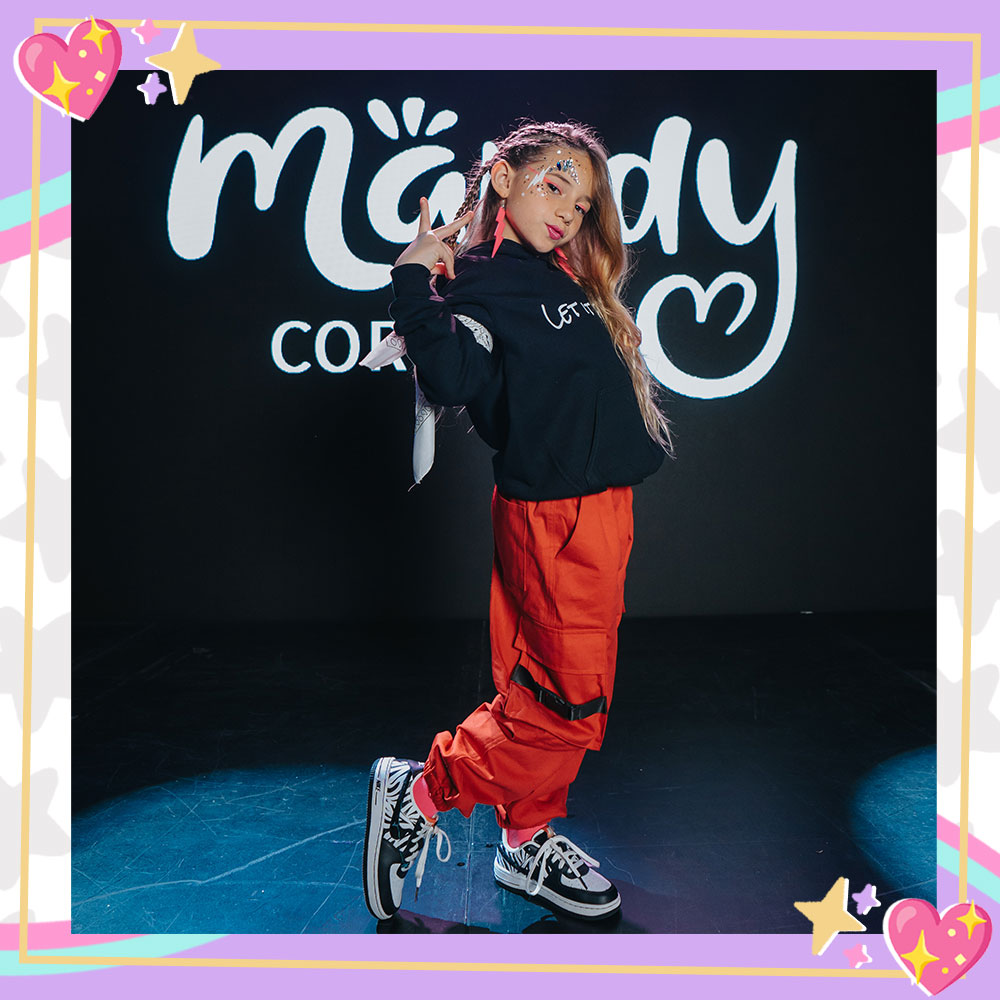 Behind the scenes photo of Mandy Corrente on the set of her Super Duper music video. She is wearing big orange cargo pants, a black sweatshirt, and big hot pink lightning bolt earrings.