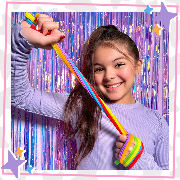 Brielle Lopez stands in front of a purple metallic streamer backdrop pulling on stretchy fidget toys. She is wearing a purple long sleeve shirt and a high ponytail.