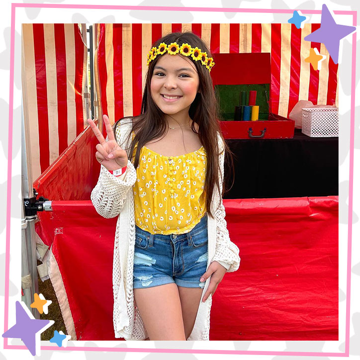 Brielle Lopez poses at a carnival. She is wearing a yellow tank top, ripped denim shorts, a long white cardigan, and a flower crown made of sunflowers. She is giving the peace sign.