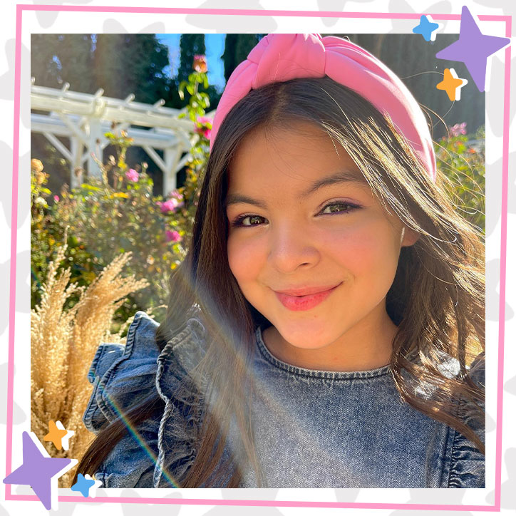 Brielle Lopez standing in a garden with a white gazebo. She is wearing a ruffled denim dress and a big pink headband.