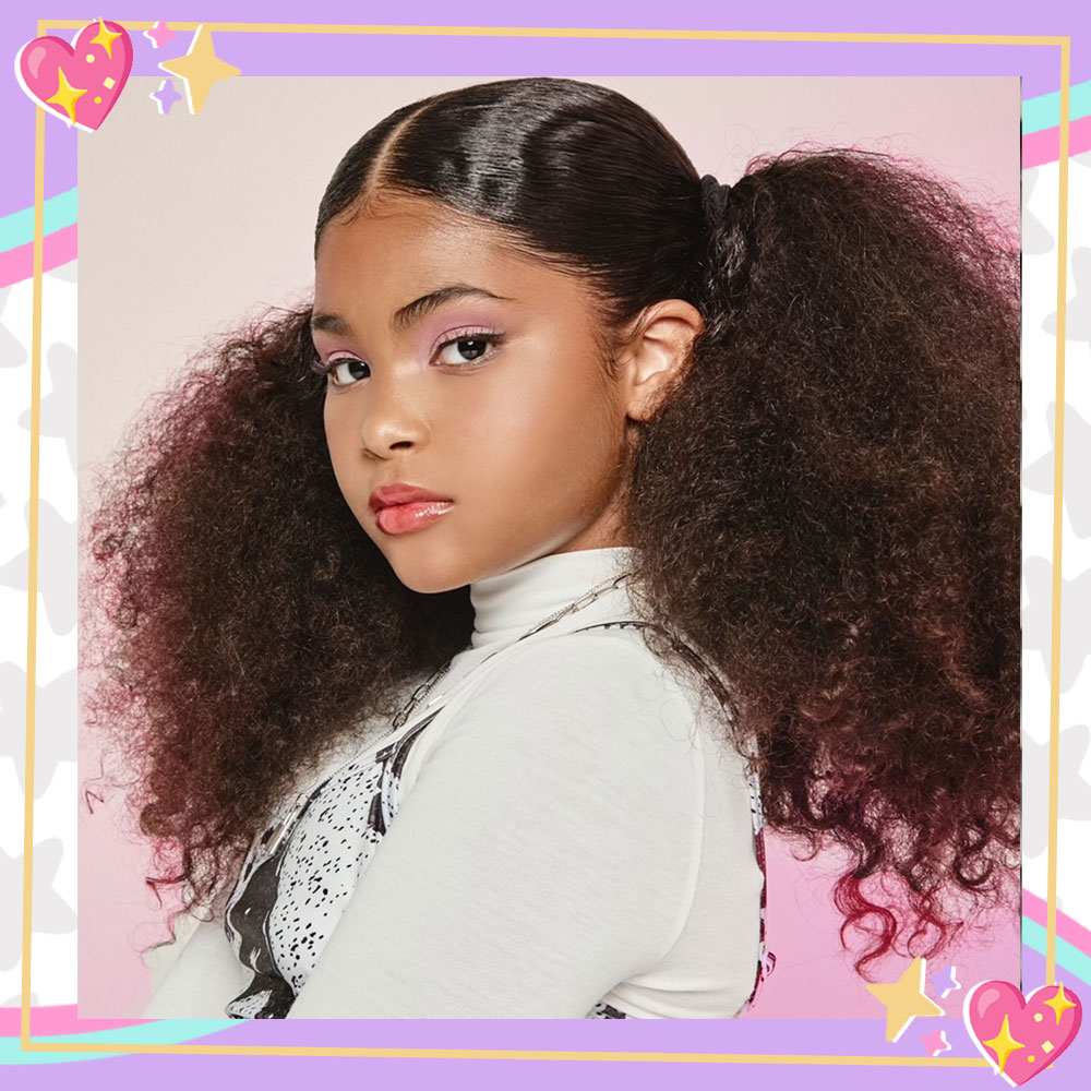 Jaidyn Triplett poses in front of a light pink backdrop. She is turned to the left, looking at the camera, arms folded. Her hair is up in big, teased curly pigtails.