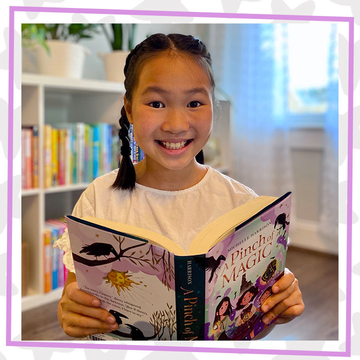 BookTuber Read With Val poses in front of a bookshelf with books organized by color. She is holding a copy of A Pinch of Magic and smiling.