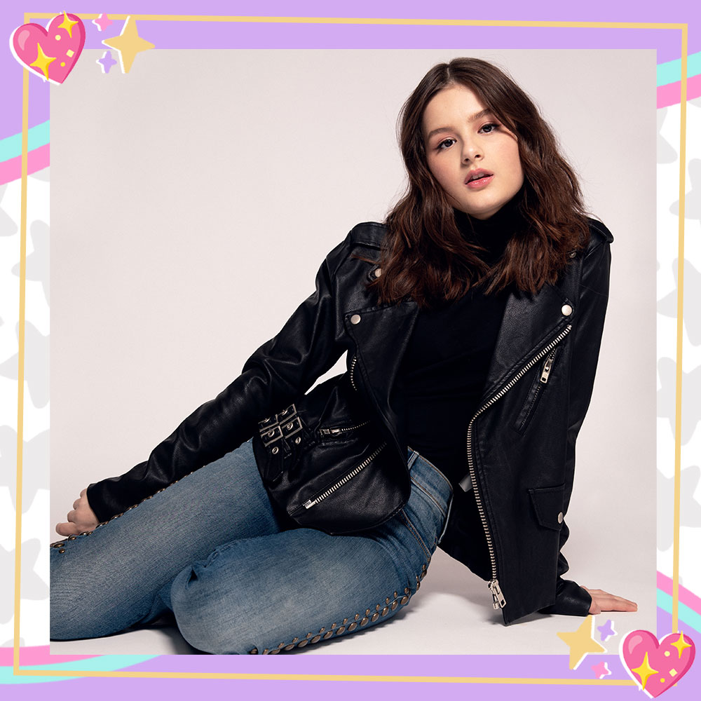 Hayley LeBlanc sits on the floor, leaning on her left arm. She is wearing a black leather jacket, black shirt, and blue jeans.