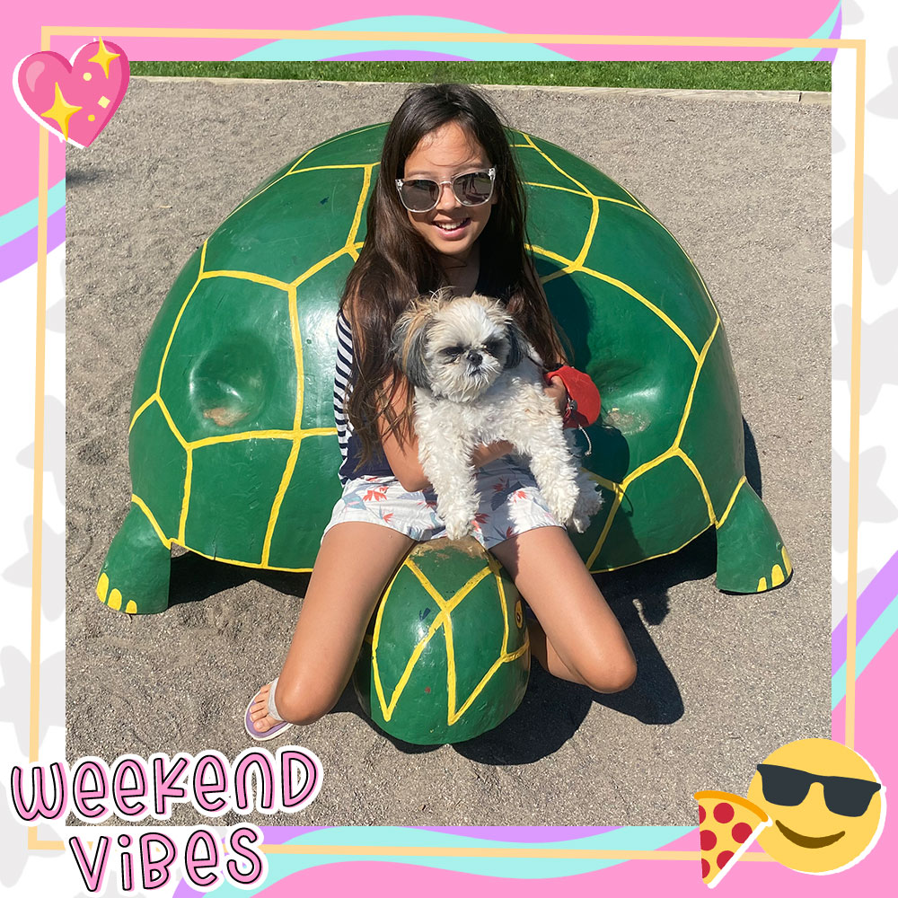 Joselyn Picard sits on a large playground turtle statue, holding a fluffy white dog in her lap. She is wearing sunglasses.