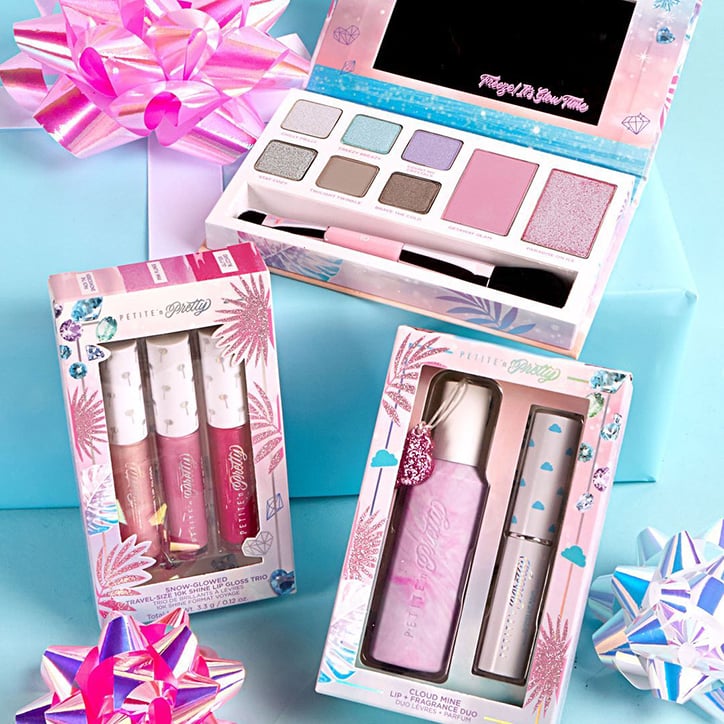Flat lay featuring the Petite 'n Pretty Paradise on Ice Makeup Collection. An eyeshadow palette, lip gloss trip, and fragrance and lip balm set are laid out on a blue table covered with metallic pink and white gift bows.